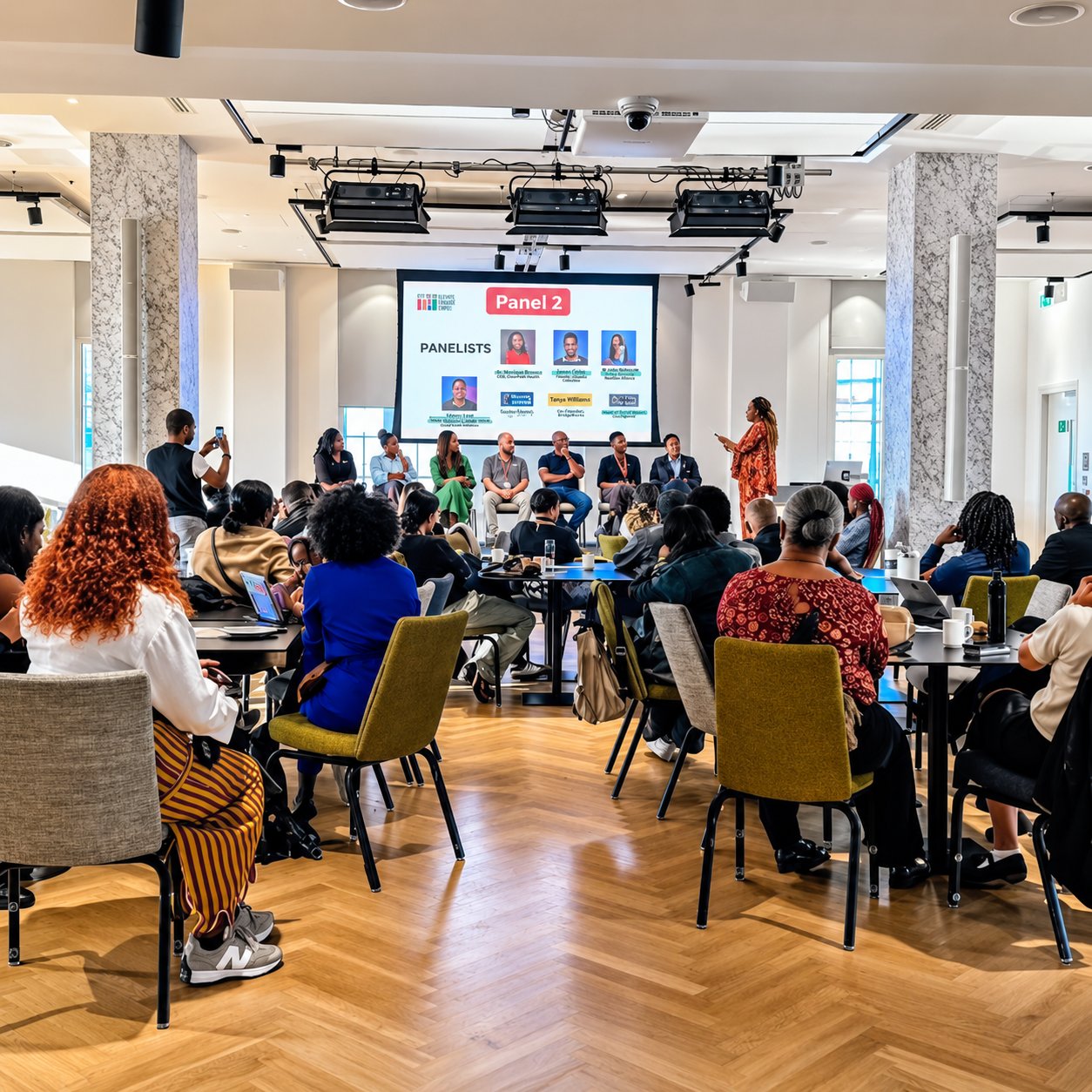 An audience of Black people seated at tables in a bright event hall listens to a panel of seven speakers, while a moderator in an orange patterned dress stands beside a screen introducing the panellists.