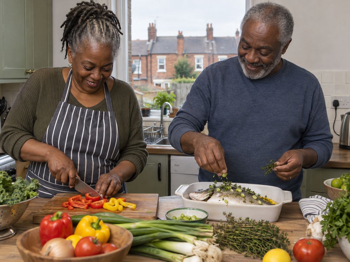 An older Black couple prepare fresh peppers, spring onions and herbs together in their kitchen, with brick terraced houses visible through the window.