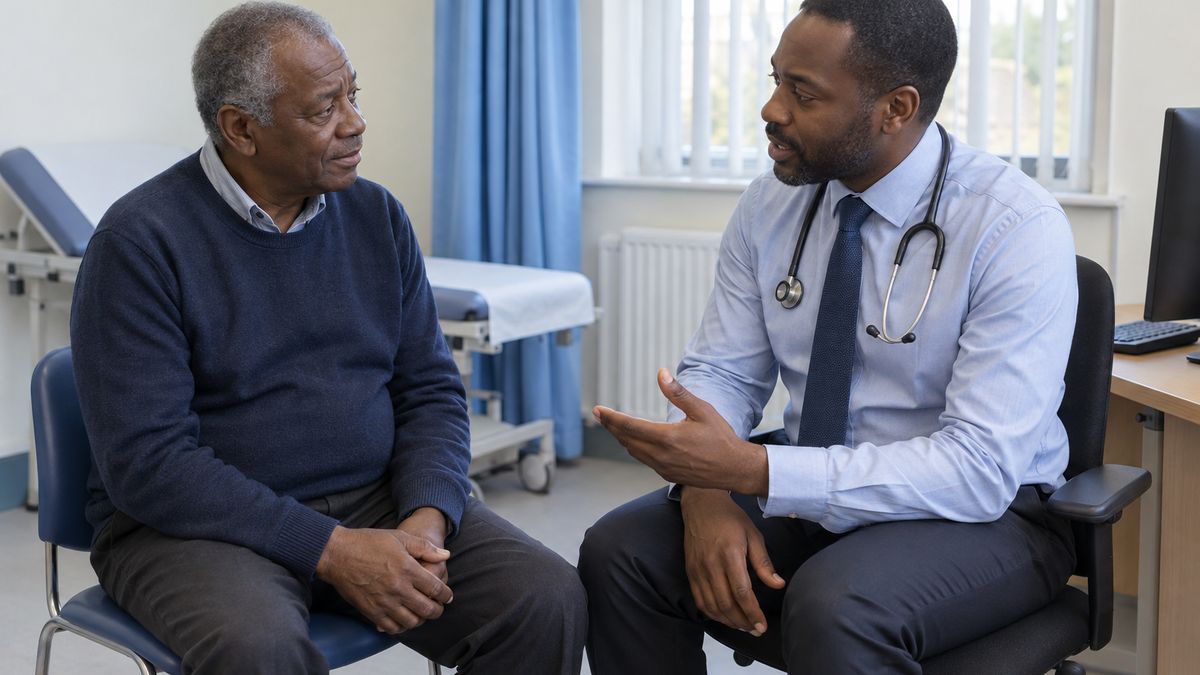 An older Black man in a navy jumper listens as a Black male doctor with a stethoscope explains something in a hospital consulting room.