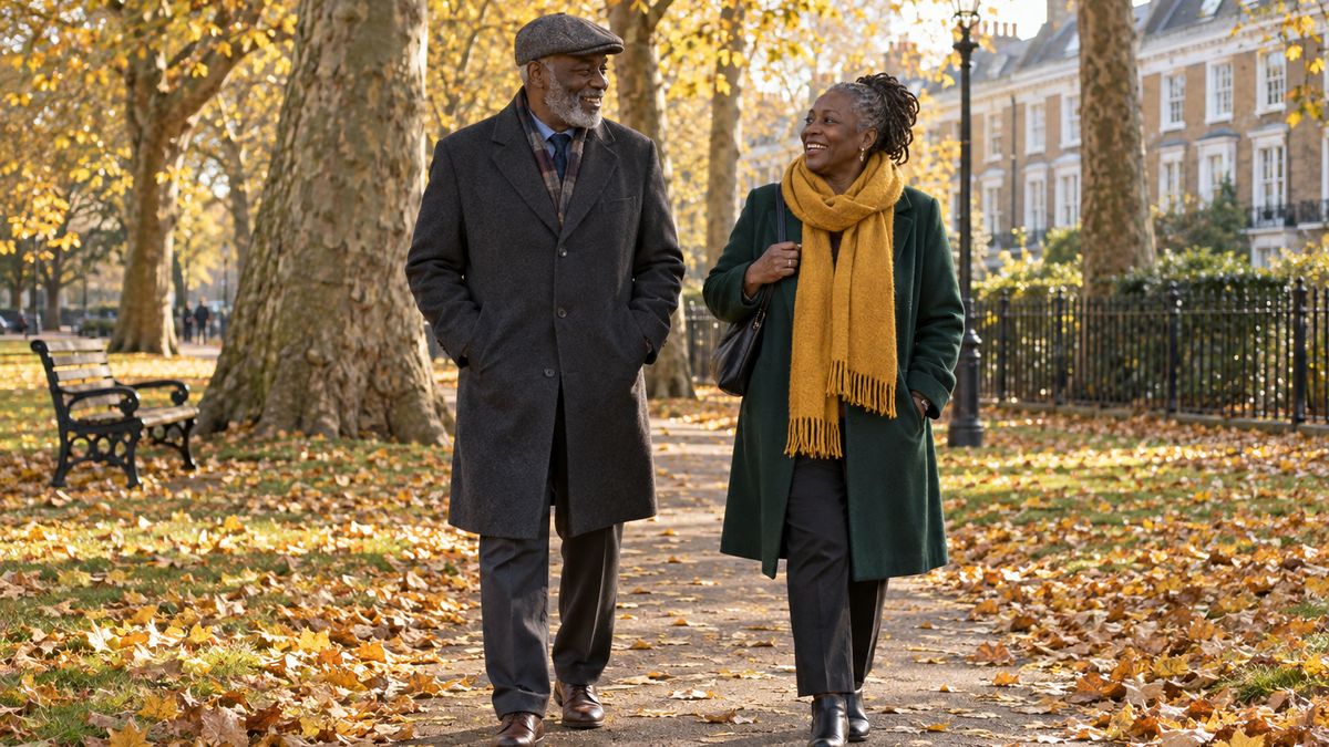 An older Black man in a flat cap and grey coat walks and laughs with a Black woman in a green coat and mustard scarf along a leaf-covered path in a London park in autumn.